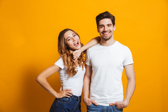 Photo of beautiful couple man and woman in basic clothing standing together and smiling at camera, isolated over yellow background