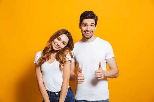 Portrait of cheerful couple man and woman 20s in basic clothing smiling and showing thumb up at camera, isolated over yellow background