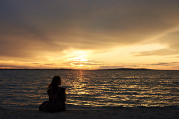 Back view silhouette of female tourist with backpack sitting alone on seashore at water edge, enjoying beautiful view of sunset on dark evening sky background. Tourism and vacations concept.