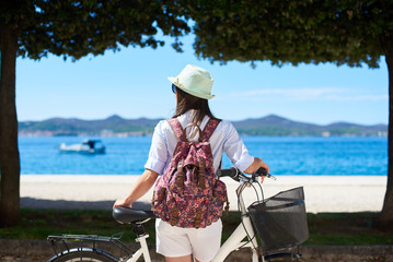 Back view female tourist in white closing, sunhat and backpack standing with bicycle on sidewalk...