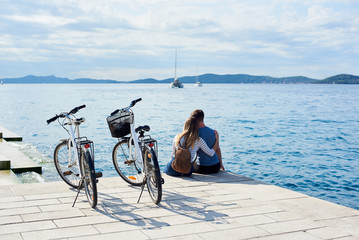 Back view of tourist couple, man and girl with backpack sitting embraced at bicycles on high paved...