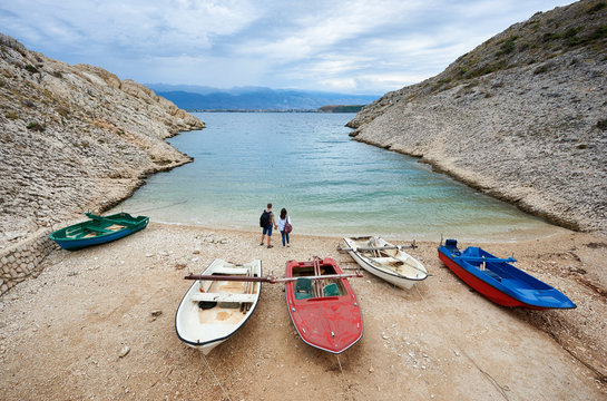Small motor boats on cozy harbor coast among high rocky shores and two young tourists, man and woman with backpacks holding hands standing at water edge. Tourism, fishing, diving, recreation concept.