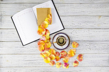 Fototapeta na wymiar a diary, a yellow envelope, petals of yellow-red roses and a cup of coffee on a saucer on a light wooden table, place for text, Flat-lay
