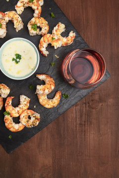 Overhead photo of plate of cooked shrimps on a dark rustic background, with a sauce and a glass of wine, with copy space