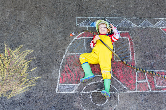 Little kid boy in fireman uniform having fun with fire truck picture drawing with colorful chalk on asphalt
