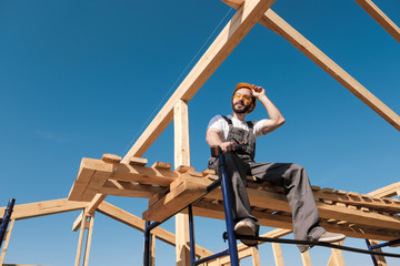 The man builder sits on the edge of the roof of the frame house, in a yellow helmet and gray overalls. The blue sky and clear sunny day. - obrazy, fototapety, plakaty