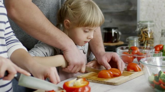 Close-up of cute little girl cutting tomato with help of father and eating one slice while cooking salad with parents