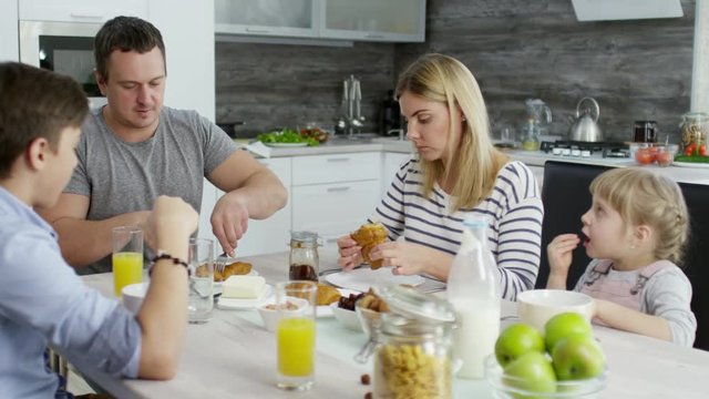 Tilt up of happy Caucasian family having breakfast at kitchen table together: parents eating croissants and kids enjoying cereals