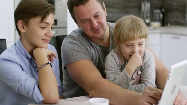 Cheerful father enjoying game on digital tablet with little daughter and teen son: family sitting at kitchen table, smiling and laughing while girl making face at front camera