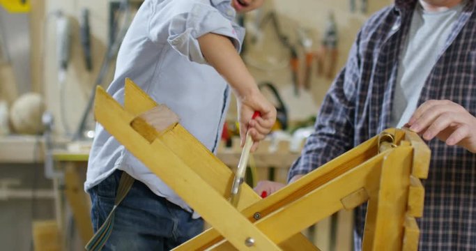 Handheld shot of happy Asian boy and his father staining wooden stool and chatting while working in garage