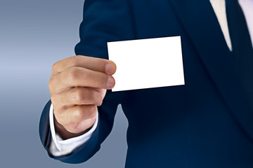 A businessman stands elegantly dressed in a blue suit and showing a business card held in hand.