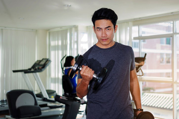 muscular asian young handsome man doing exercises with dumbbell for good healthy in fitness gym at morning with people background, bodybuilder, lifestyle, exercise workout and sport training concept