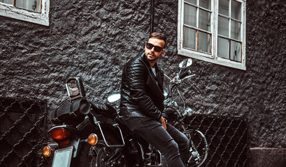 Fototapeta na wymiar Fashionable biker dressed in a black leather jacket and jeans sitting on his retro motorcycle on an old Europe street.