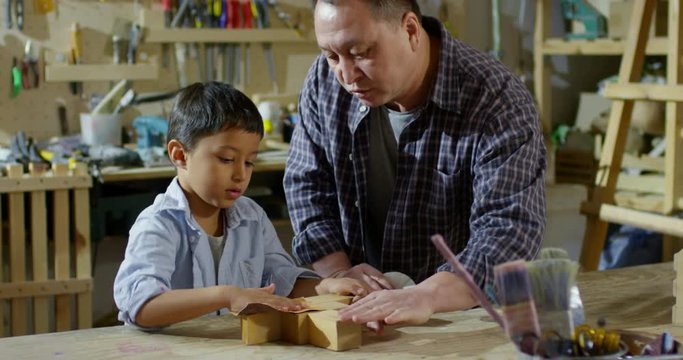 Tilt up of middle-aged father in plaid shirt giving advice to cute little son sanding wooden toy with sandpaper in carpentry workshop