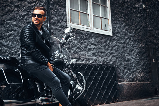 Fashionable biker dressed in a black leather jacket and jeans sitting on his retro motorcycle on an old Europe street.