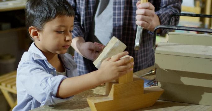 Handheld shot of unrecognizable father in plaid shirt using chisel and mallet on wooden toy while teaching his little son how to work with wood