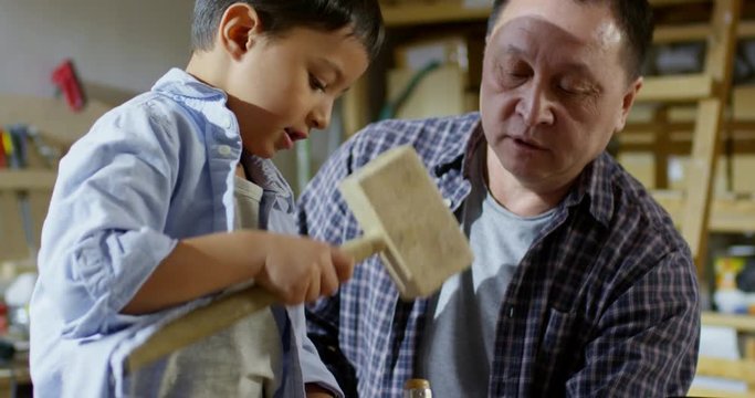 Medium shot with tilt down of middle-aged Asian man teaching little son how to use chisel and mallet while spending time with him in carpentry workshop making wooden toy