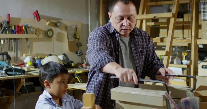 Handheld shot of middle-aged Asian man in plaid shirt opening tool box and showing chisels to cute little boy sitting at workbench and looking at unfinished wooden toy