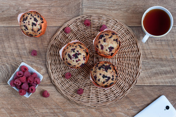 Fresh muffins with raspberry, cup of tea and smartphone are lying on a wooden desk.