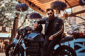 Foto op Plexiglas Stylish fashionable biker in sunglasses dressed in a black leather jacket, sitting on his custom-made retro motorcycle near terrace of cafe. © Fxquadro