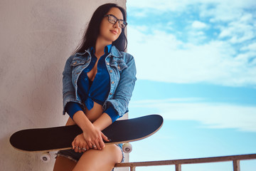 Portrait of a sexy brunette girl wearing short shorts and jeans jacket holds a skateboard while...