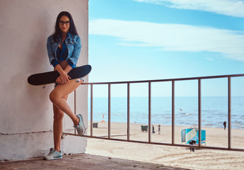 Seductive hot brunette girl wearing short shorts and jeans jacket holds a skateboard while standing...