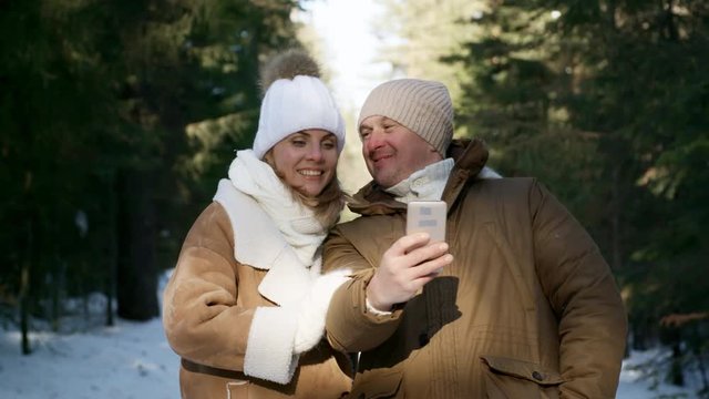 Loving couple having fun on sunny day in winter forest and taking selfie together using smartphone