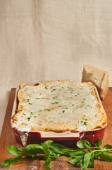 Top view, medium distance of an artisan, freshly baked, homemade lasagna in a red baking dish cooling on a wood serving board with a sprig of fresh parsley