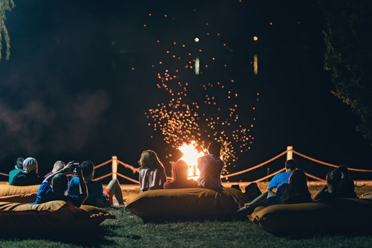 Group of people near camp fire with campfire song and campfire meals playing campfire games and eating campfire grill, telling campfire stories near the fire with wood, flames in the nature at night. - Powered by Adobe