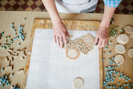 Top view of unrecognizable craftsman laying out shaped marble tiles in pattern while creating mosaic in traditional crafting workshop