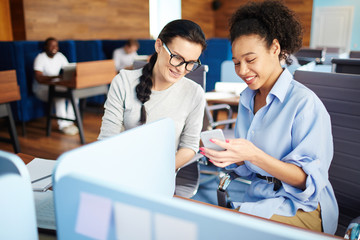 Young smiling businesswoman sitting together with her colleague and showing something on her smart phone at office
