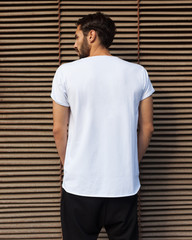 Fashion street youth. A man in a white T-shirt posing with his back to the camera on a dark blue metallic background in the summer evening. Cover. Back.