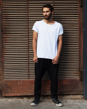 A bearded hipster in a white T-shirt, black pants and sneakers posing against a rusty metal wall.