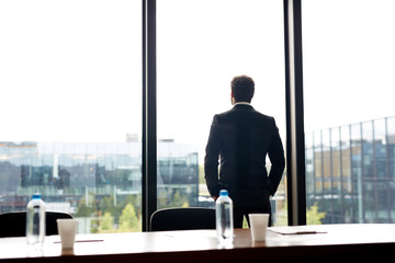 Fototapeta premium Contemplative businessman in formal suit holding hand in pockets and looking out panoramic window while waiting for conference in meeting room