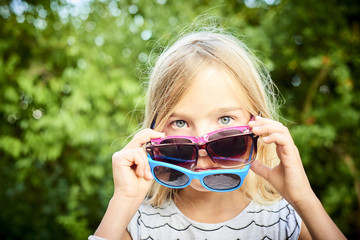 Child cute girl having several sunglasses on her face and doing fun during a hot summer day. Greenery background