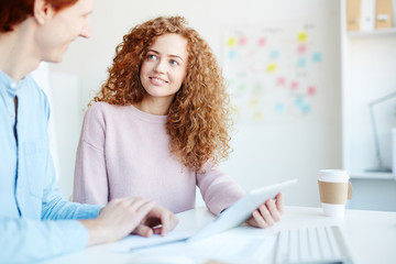 Cheerful positive confident young colleagues in casual clothing using tablet while analyzing information together and discussing it in workplace