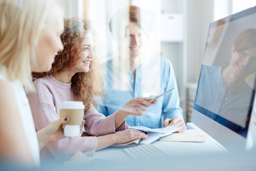 Obraz premium Group of positive young creative designers in casual clothing discussing project in office behind glass: cheerful curly-haired lady pointing at computer monitor while offering idea