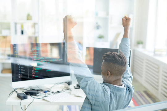 Ecstatic young African-American software developer raising hands in excitement while achieving success in web development, he sitting in front of computer behind glassy wall