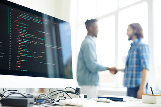 Hipster programmer and African entrepreneur making handshake in background, focus on computer monitor with programming code and messy cables on table