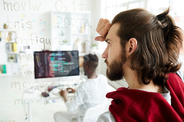 Serious confused hipster coder with pony tail looking at computer text while brainstorming about...