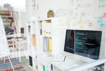 Coders workplace: glassy wall with script language, desktop computer with code on table, marketing board behind computer, hipster man working in background