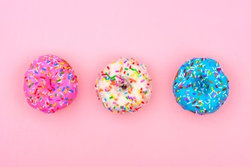 Three assorted donuts with pastel colored icing and sprinkles against a soft pink background. Minimal concept.