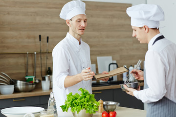 Confident skilled chef in hat holding notepad and explaining recipe while teaching intern with...