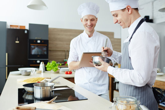 Cheerful optimistic handsome young chef satisfied with students work writing in sketchpad and laughing while talking to intern in apron mixing food in bowl in commercial kitchen.