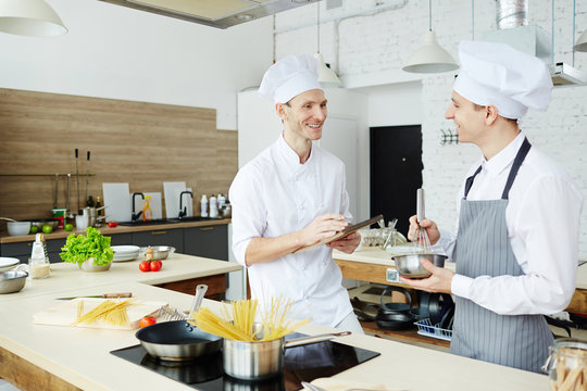 Happy skilled young cook in chefs uniform making notes while talking to intern at cooking class, smiling man in apron using whisk while whipping cream