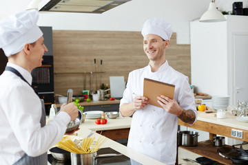 Positive pleased handsome young chef with tattoo on arm making notes in sketchpad while testing intern cook at class
