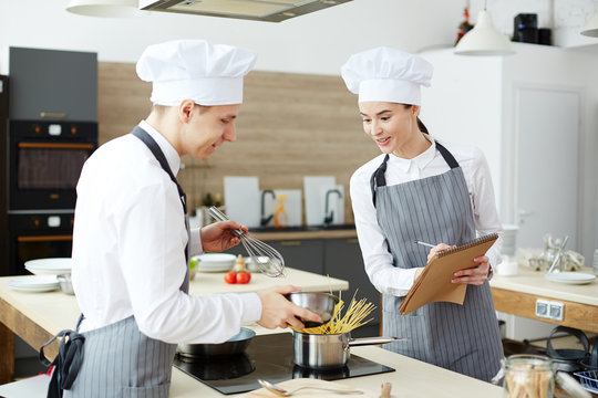 Surprised positive female cooking expert in uniform controlling young cook and checking whipped cream in bowl while writing comments in notepad in restaurant kitchen