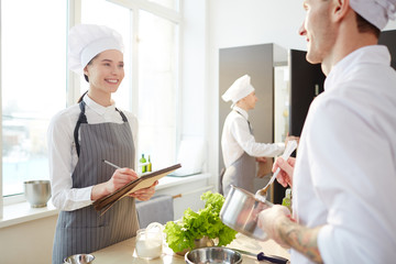 Happy positive student girl in apron knowing new recipe at cooking class and making notes in...