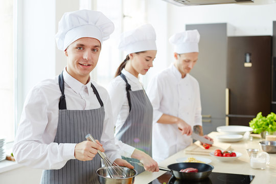 Smiling handsome young cook in apron and chefs hat whipping ingredients in bowl and looking at camera while his colleagues cooking in background