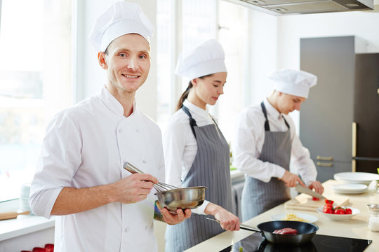 Happy confident professional chef in white uniform using whisk for whipping and looking at camera while working in commercial kitchen